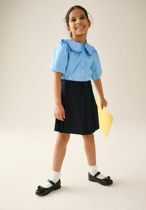 Lächelndes Mädchen in einer blauen Bluse mit Rüschenkragen, schwarzem Rock, weißen Socken und schwarzen Schuhen, das eine gelbe Mappe hält, stehend auf hellem Hintergrund.
