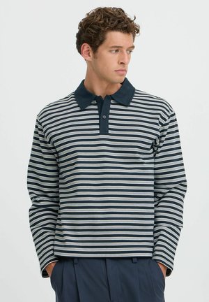 Jonge man die een marineblauw-wit gestreept poloshirt met lange mouwen en marineblauwe broek draagt, kijkt opzij met handen in de zakken.