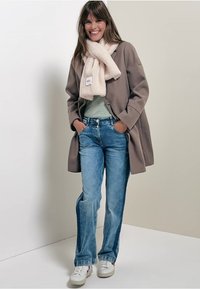 Beige wollen jas over een lichtgroene sweater, gecombineerd met blauwe jeans en een pluizige roze sjaal. Witte sneakers met rode accenten maken de look compleet.