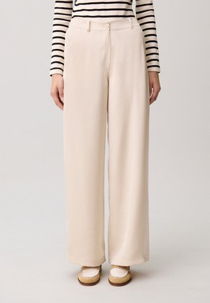 Mujer vestida con pantalones anchos beige, mocasines crema y marrón claro, y una camiseta de manga larga a rayas blancas y negras, de pie sobre un fondo claro y liso.