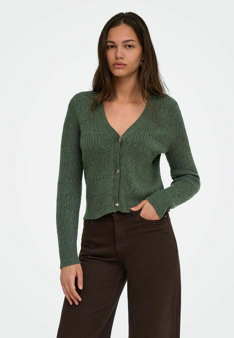 Jeune femme aux longs cheveux foncés portant un pull vert boutonné et un pantalon marron foncé, une main dans la poche.