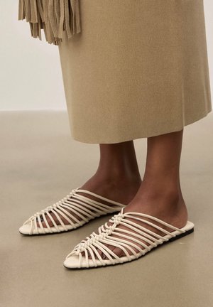Pieds portant des sandales plates beiges à brides, assorties à une jupe beige ornée de franges au bas.