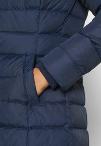 Dłoń osoby częściowo wsunięta do bocznej kieszeni granatowej pikowanej zimowej kurtki z widocznym ściągaczem na rękawie.