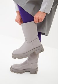 Botas grises hasta la rodilla con acabado mate. Suela de goma gruesa y diseño recto. Llevadas con mallas moradas y una prenda gris.