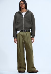 Gray zip-up hoodie with large front pockets, paired with loose olive green trousers. Black belt and accessories complete the outfit.