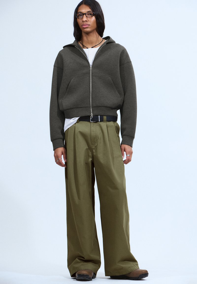 Gray zip-up hoodie with large front pockets, paired with loose olive green trousers. Black belt and accessories complete the outfit.