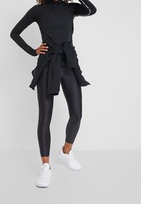 Černý dlouhý rukáv s vysokým límcem, doplněný lesklými černými legínami a bílými sportovními botami. Detaily zahrnují svetr uvázaný kolem pasu.