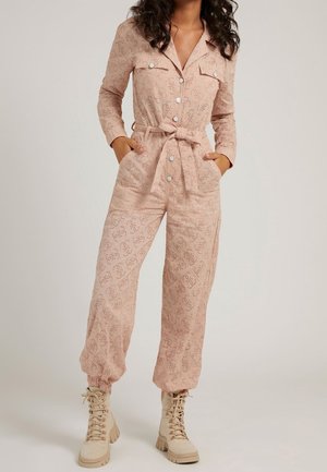 Femme portant une combinaison beige en œillets avec manches longues, boutons, ceinture et poches, associée à des bottines beiges à lacets.