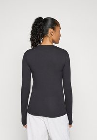 Frau mit lockigem Pferdeschwanz, die ein enges, langärmliges schwarzes Shirt und hellgraue Hose trägt, von hinten vor einem schlichten Hintergrund betrachtet.