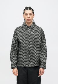 Jaunas žmogus pintais plaukais dėvi juodą sagotu švarką, padengtą pasikartojančiu pilku lapų raštu, ir juodas kelnes su baltais dryžiais.