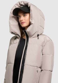 Helle beige Daunenjacke mit großer Kapuze, ausgestattet mit einem durchgehenden Reißverschluss und schwarzen Akzenten, kombiniert mit einer schwarzen Mütze. Glatte Textur, gepolstertes Design.