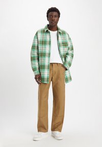 Camisa de franela a cuadros verde y blanca sobre una camiseta blanca, combinada con pantalones anchos color topo y zapatillas blancas. Diseño sencillo, look casual.