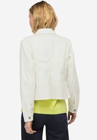 Weiße, kurz geschnittene Jeansjacke mit einem taillierten Kragen, geknöpften Manschetten und Rückennähten, kombiniert mit einem leuchtend gelben Oberteil darunter.