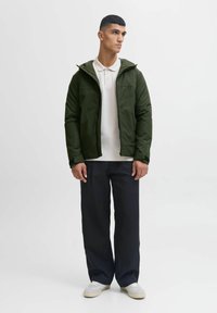 Giacca isolante verde con cappuccio, zip nera e polsini elasticizzati, abbinata a una polo bianca e pantaloni neri larghi. Scarpe leggere.
