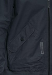 Marineblauwe bomberjack met een zijzak met knoopsluiting, ritssluiting aan de voorkant, geribde manchetten en een label met de tekst "Gemaakt voor alle omstandigheden."