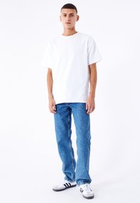 Ung mand står i blå jeans, hvid t-shirt og hvide sneakers med sorte striber foran en plain hvid baggrund.