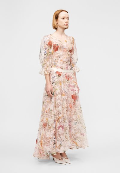 Γυναίκα που φοράει ένα διάφανο, μακρύ φόρεμα με λουλούδια και φουσκωτά μανίκια, καθώς και λευκά τακούνια με μυτερή μύτη, στέκεται μπροστά σε ένα απλό ανοιχτό φόντο.