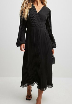 Woman wearing a long-sleeve, pleated black midi dress with a V-neck, paired with black heeled sandals and holding a black handbag.