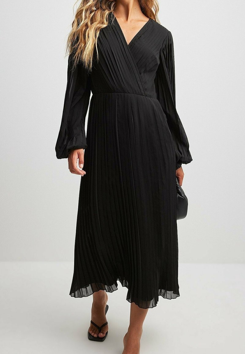 Femme portant une robe midi noire plissée à manches longues avec un col en V, associée à des sandales à talons noires et tenant un sac à main noir.
