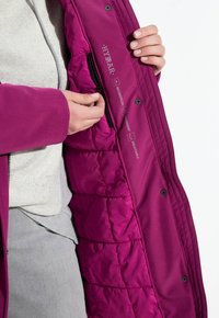 Person wearing gray pants and cream hoodie showing a magenta HYBRAR jacket interior with zippered pocket and product feature labels.