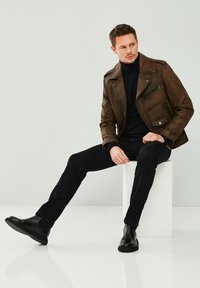 Veste de moto en cuir marron avec poches zippées, portée sur un col roulé noir, associée à un pantalon noir et des bottines, assise sur un bloc blanc.