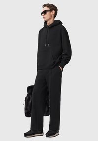 Czarny sweatshirt z kieszenią z przodu i sznurkiem, w połączeniu z pasującymi czarnymi dresami i czarnymi trampkami. Tło jest jasnoszare.