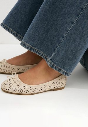 Pieds portant des ballerines beiges perforées avec de petits nœuds, associées à un jean denim bleu à jambes larges.