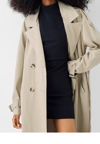 Beige trenchcoat med stora knappar, med en svart tajt klänning under. Kåpan har breda slag och bälte vid ärmarna. Slät tygtextur.