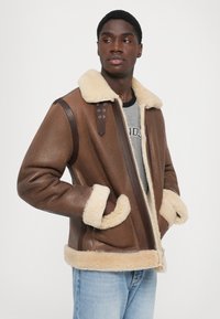 Veste en shearling brun avec un col et des poignets en fourrure crême, dotée d'une fermeture éclair à l'avant et d'accents en cuir brun sur les épaules.