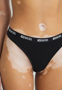 Svart stringtrosor med en vit "DIESEL"-logo på midjebandet, med slät tyg och låg midja.