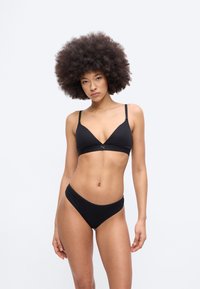 Žena s veľkými kučeravými afro vlasmi modeluje čiernu podprsenku a nohavičky so štíhlym strihom na jednoduchom svetlosivom pozadí.