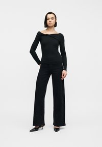 Άτομο με κοντά σκούρα μαλλιά που φοράει εφαρμοστό μαύρο τοπ χωρίς ώμους, μαύρο παντελόνι με φαρδιά πόδια και μαύρες μυτερές γόβες με τακούνι kitten, στέκεται με αυτοπεποίθηση.