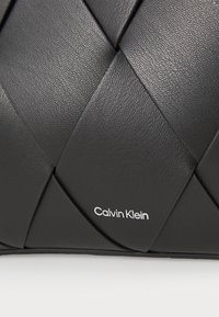 Crna kožna torba s tkanom teksturom i elegantnim dizajnom. Ima srebrni "Calvin Klein" logotip s jedne strane.