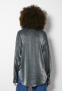 Långärmad, metallisk silverfärgad skjorta med en avslappnad passform, mjuk textur, knappärmar och en rundad nederkant, i kombination med blå byxor.