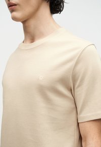 Κοντινό πλάνο ενός ατόμου που φοράει απλό μπεζ κοντομάνικο μπλουζάκι με μικρό, διακριτικό λογότυπο στο στήθος, σε λευκό φόντο.