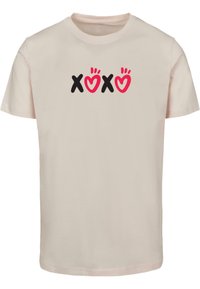 Lysbeige bomulds-t-shirt med rund halsudskæring og kortærmer, der viser et grafisk print af "XOXO" i sorte og lyserøde farver.