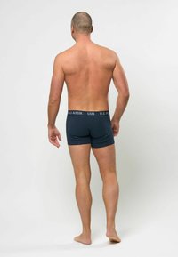 Marineblå bomullsbokser med en kontrasterende midjebånd merket "U.S. POLO ASSN." Modellen vist fra baksiden, barføtt, uten skjorte.