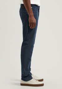 Pantalones azul marino de algodón con corte ajustado. Presentan cinco bolsillos, costuras visibles y un parche con el logo en la cintura.