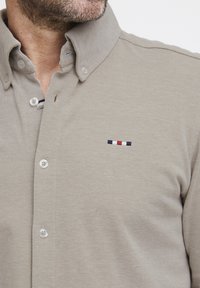Chemise beige à boutons avec un tissu texturé, présentant un col classique et un logo brodé en marine, blanc et rouge sur la poche poitrine.