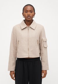 Beige kort jacka med dragkedja, stor krage, två framfickor med dragkedja och en sidoficka med guld detaljer.