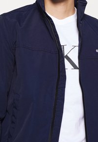 Námořnická bunda na zip s stojáčkovým límcem, s detaily švů a dvěma bočními kapsami; nošená přes bílou košili s černým potiskem.