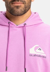 Svetloružová mikina s kapucňou a šnúrkami, na ktorej je biele logo Quiksilver. Materiál vyzerá mäkko, má hladkú textúru a voľný strih.