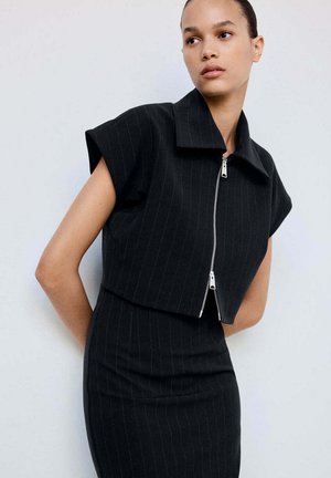 Vrouw draagt een zwart pinstripe cropped top met korte mouwen en een bijpassende hoge taille rok, staand tegen een effen lichte achtergrond.