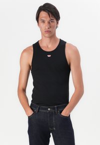 Camiseta sin mangas negra con cuello redondo, fabricada en un tejido suave, que presenta un pequeño logo rojo en el pecho, combinada con unos jeans de mezclilla oscura.