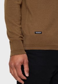Bruine lange mouwen trui met geribbelde manchetten en zoom. Zwarte "THREADBARE" label onderaan. Hand rustend op denimstof.