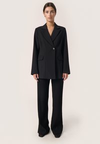 Schwarz maßgeschneiderter Blazer mit einem Knopf, Revers mit Kerb, und vordere Taschen, kombiniert mit schwarz gerippten, ausgestellten Hosen. Glatte Textur.