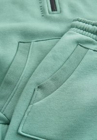 Hellgrüner Sweatshirtstoff mit auffälligen Nähten, ausgestattet mit zwei schrägen Taschen und einer kleinen mit Reißverschluss versehenen Tasche auf der Brust. Glatte Textur.
