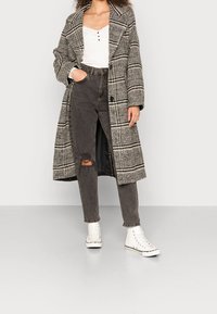 Kostkovaný kabát v šedé a černé látce, s velkým límcem, předními knoflíky a kapsami, přes šedé roztrhané džíny a bílé vysoké tenisky.