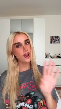 Grijze oversized t-shirt met een kleurrijk grafisch ontwerp van een appel. Lang, steil blond haar, met grote zilveren oorbellen.