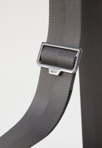 Šedý textilní pásek s texturovaným povrchem, zdobený elegantní stříbrnou kovovou přezkou a detaily loga, představující jednoduchý a funkční design.